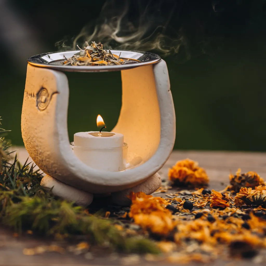 Keramik-Räuchergefäß mit brennendem Teelicht und glimmendem Räucherwerk, umgeben von Kräutern.