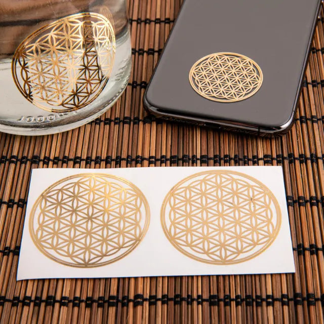 Zwei goldene 3D-Aufkleber 'Blume des Lebens' auf einem Blatt, einer auf Glas, einer auf Smartphone.