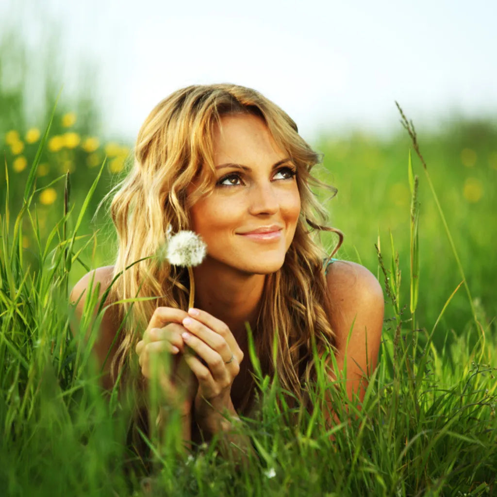 Frau im Gras mit Pusteblume, symbolisiert Lebensfreude, Leichtigkeit und Naturverbundenheit.