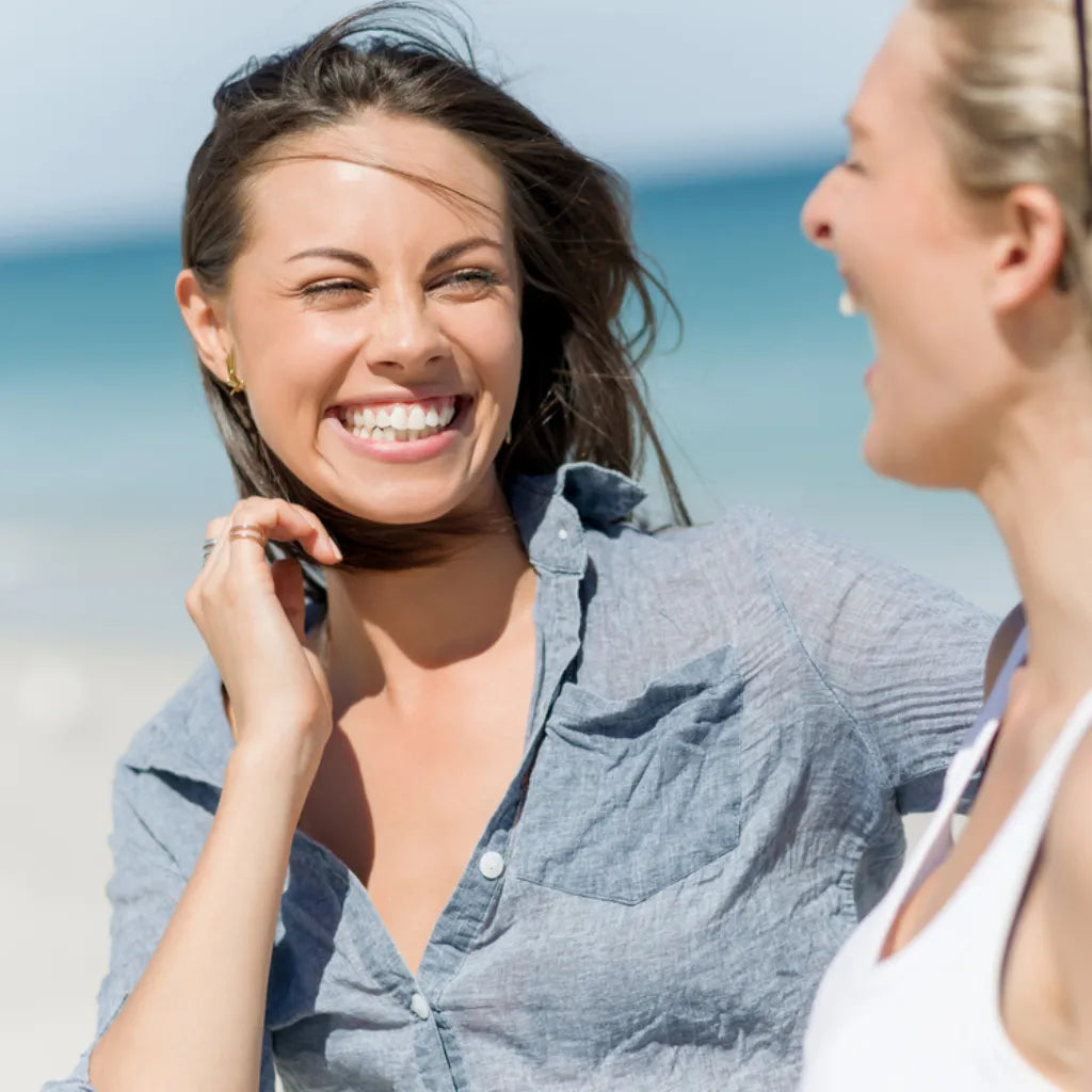Zwei Frauen lachen am Strand, symbolisieren Freude, inneren Frieden & positive Anziehungskraft im Leben.
