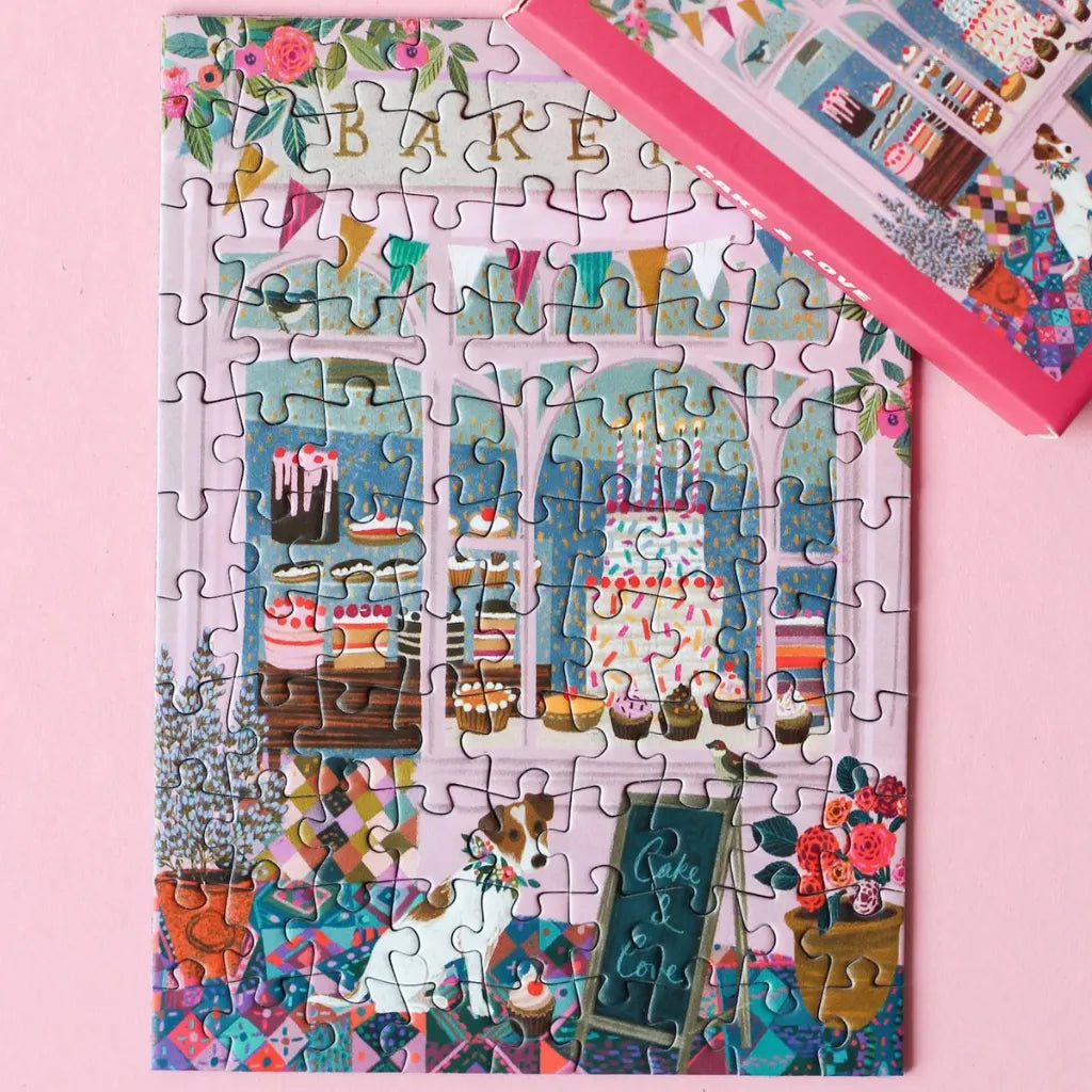 Das vollständige 99 Teile Piecely Minipuzzle "Cake & Love" neben seiner pinken Verpackung. Präsentiert das süße Bäckerei-Motiv mit Hund, Schaufenster mit Kuchen und floraler Dekoration.