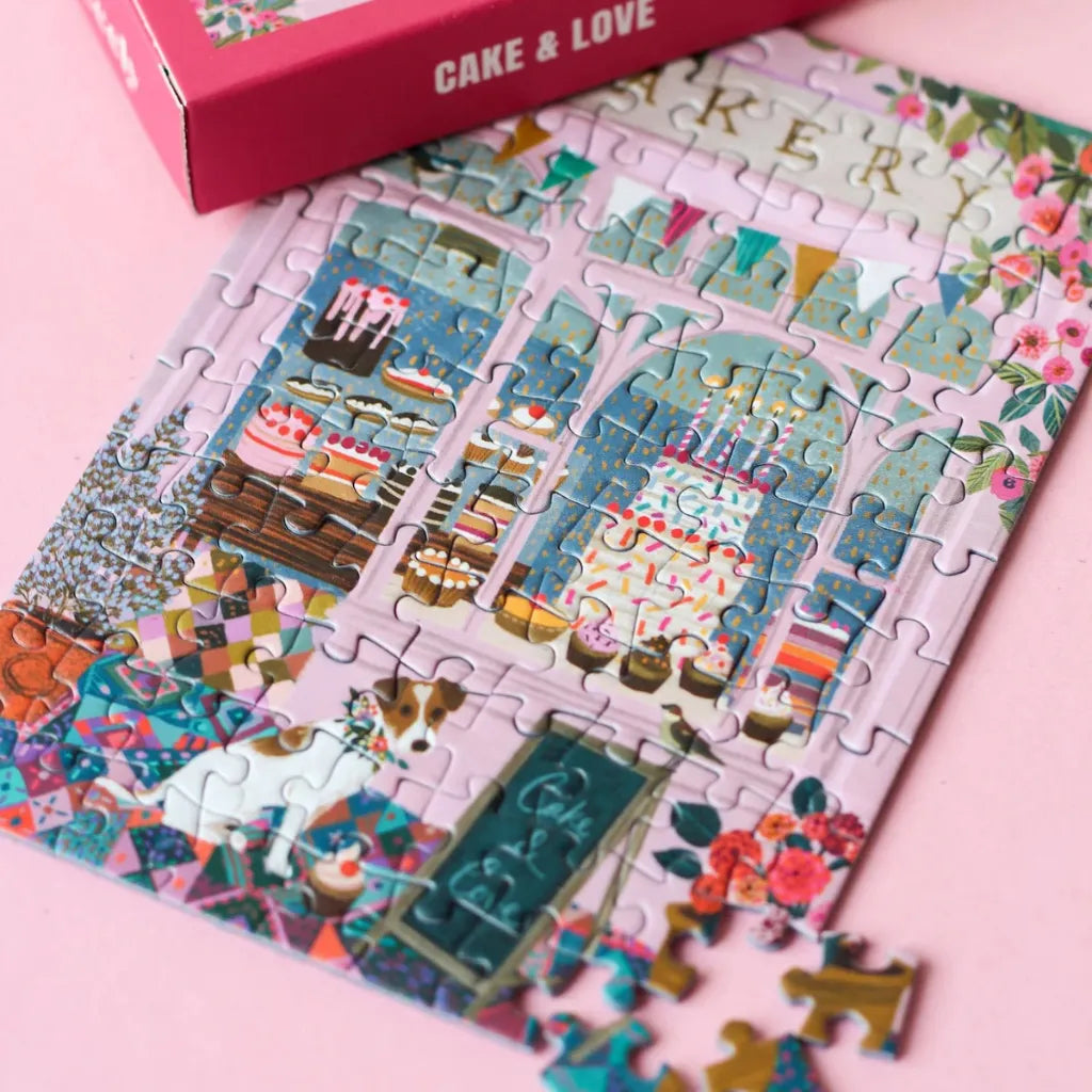 Teilweise fertig gelegtes 99 Teile Piecely Minipuzzle "Cake & Love" mit der rosa Verpackung und einzelnen Puzzleteilen. Nahaufnahme des Bäckerei-Motivs auf pinkem Hintergrund.