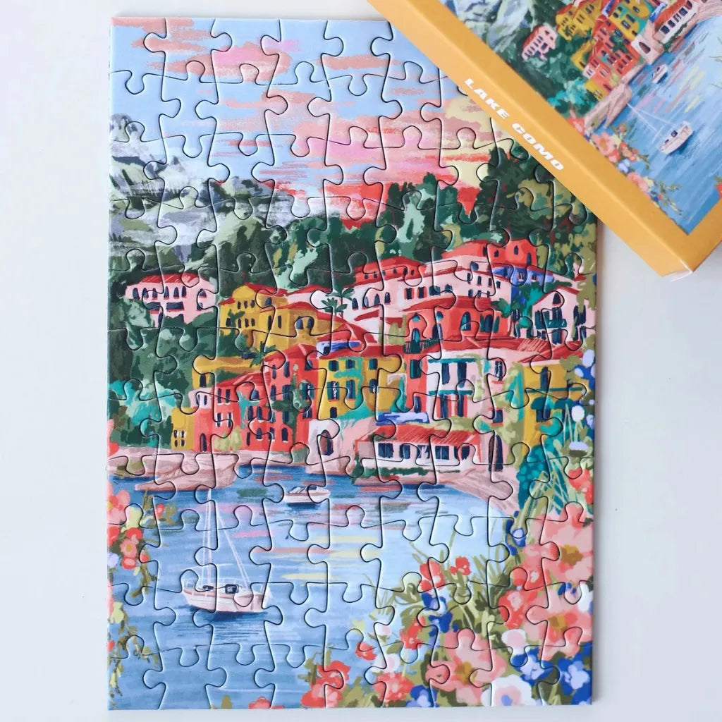 Das fertig gelegte 99 Teile Piecely Minipuzzle "Lake Como" liegt neben der gelben Verpackung. Zeigt die charmante Szene des Comer Sees mit seinen charakteristischen Gebäuden und der umliegenden Natur.