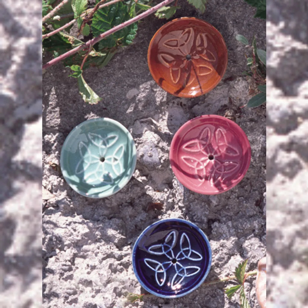 Vier runde Keramik Räucherstäbchenhalter in den Farben Rotbraun, Hellgrün, Pink und Dunkelblau mit harmonischem Knotenmuster, auf einer rauen Steinoberfläche mit Pflanzen – ideal für entspannte Momente.