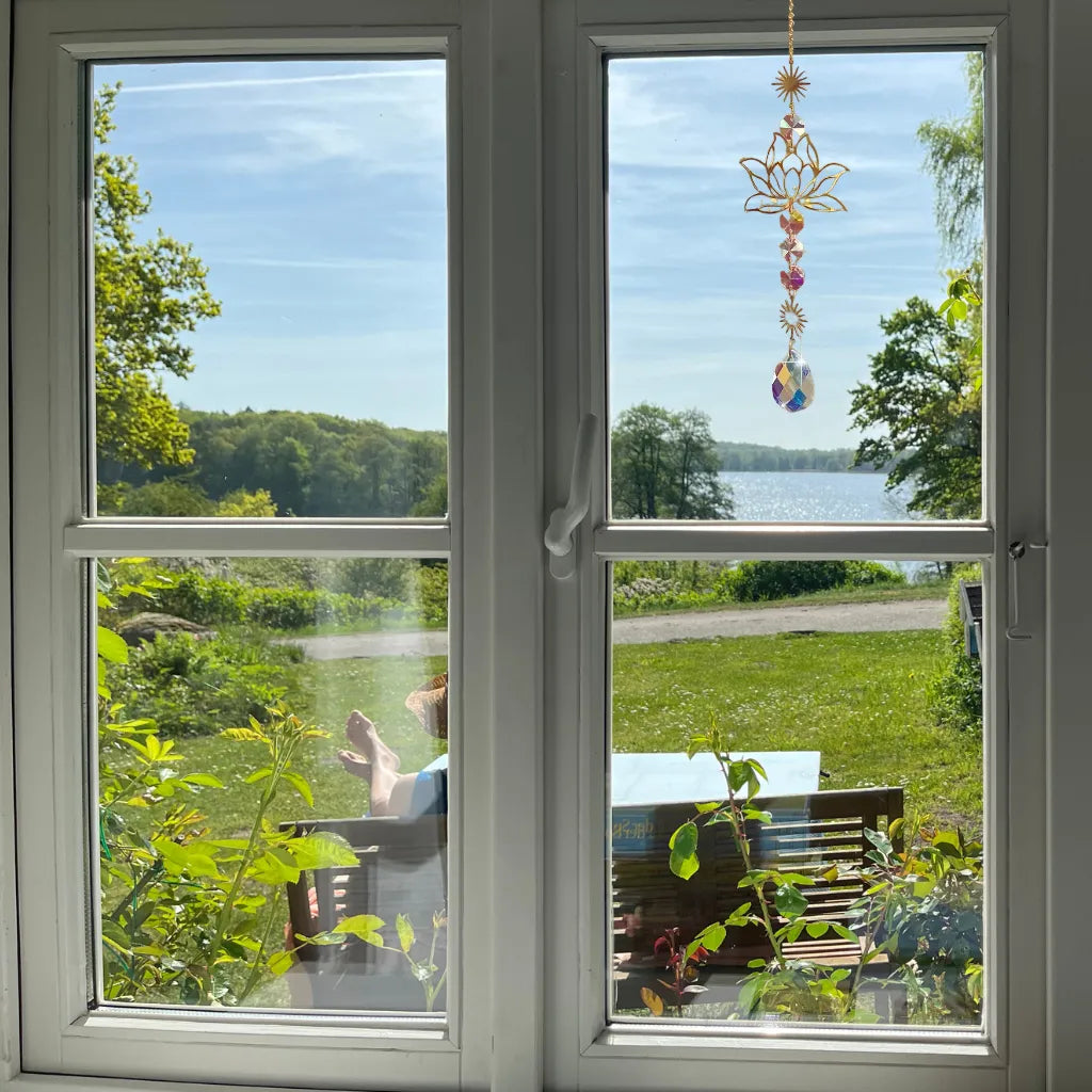 Elegante Sonnenfänger Kette mit goldenem Lotusmotiv am Fenster mit Seeblick, bringt Ruhe und positive Energie in den Raum.