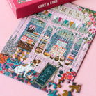 Teilweise fertig gelegtes 99 Teile Piecely Minipuzzle "Cake & Love" mit der rosa Verpackung und einzelnen Puzzleteilen. Nahaufnahme des Bäckerei-Motivs auf pinkem Hintergrund.