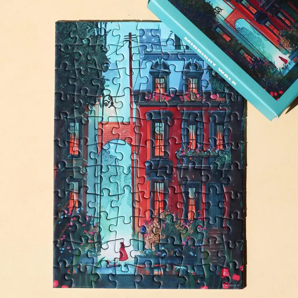 Das fertig gelegte 99 Teile Piecely Minipuzzle "Midnight Walk" neben der dazu passenden, türkisfarbenen Verpackung. Eine atmosphärische Ansicht der nächtlichen Stadt mit Gebäuden und Grün.