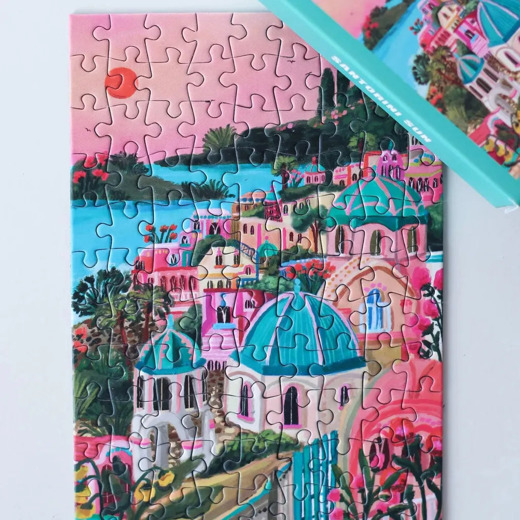 Das fertige 99 Teile Piecely Minipuzzle "Santorini Sun" liegt neben der türkisfarbenen Verpackung. Eine malerische Ansicht der ikonischen Architektur und lebhaften Landschaft Santorinis.