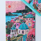 Das fertige 99 Teile Piecely Minipuzzle "Santorini Sun" liegt neben der türkisfarbenen Verpackung. Eine malerische Ansicht der ikonischen Architektur und lebhaften Landschaft Santorinis.